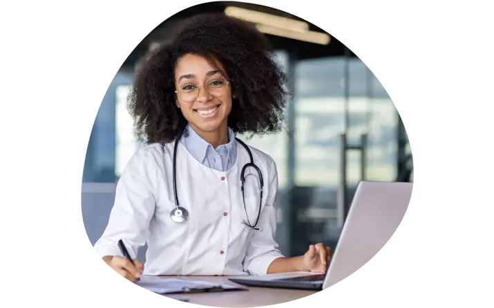 Image of a doctor sitting behind a desk with a laptop and notes on it whilst smiling